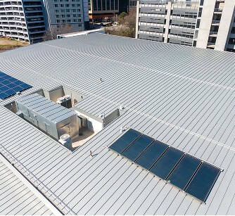Aerial of a commercial flat metal roof with solar panels, HVAC units, and nearby office buildings, ideal for flat roof restoration and commercial roof repair.