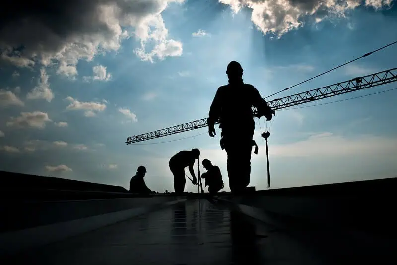 Silhouetted workers repair a commercial flat roof under cloudy skies with a crane.