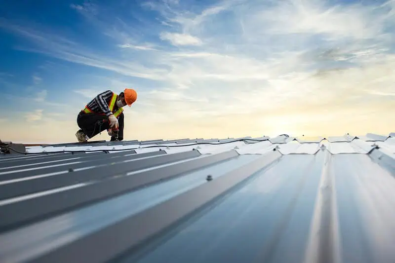 A worker in safety gear inspects a commercial metal flat roof at sunset for repair.