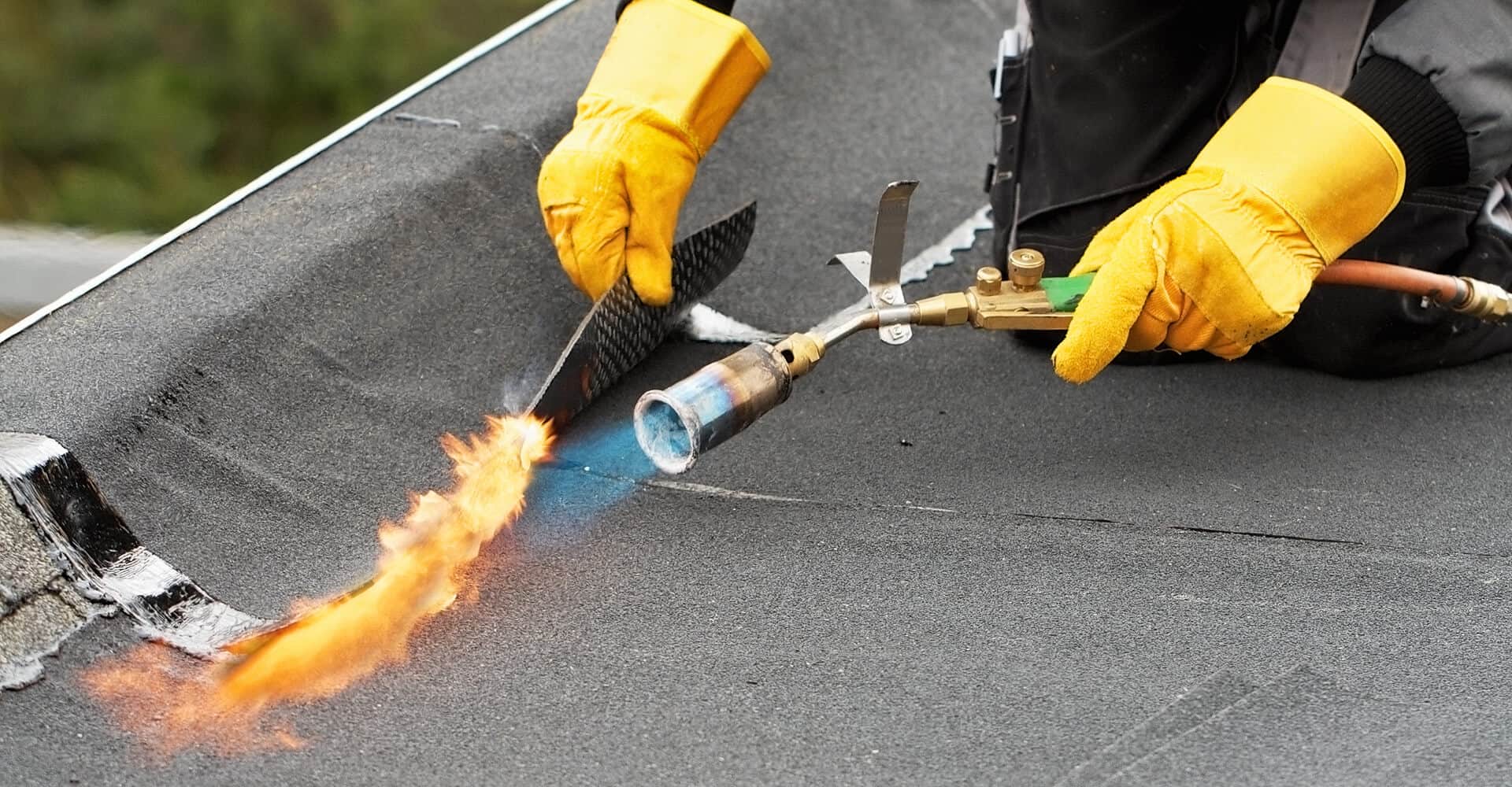 A worker with yellow gloves uses a blowtorch for commercial flat roof repair.