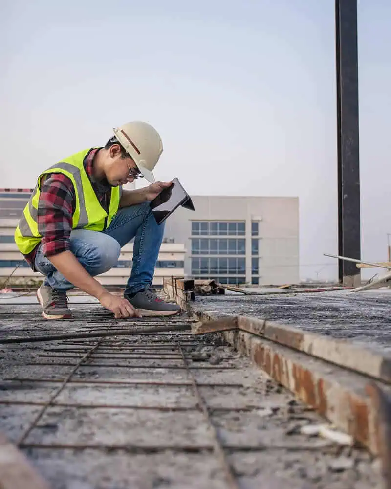 A worker inspects rebar on a commercial flat roof while reviewing repair plans on a tablet.