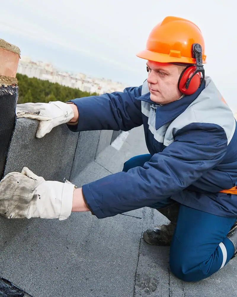 A worker in safety gear repairs a commercial flat roof near a chimney vent.